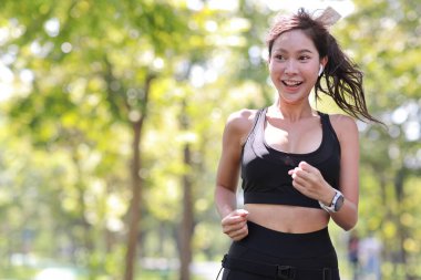 Güzel Asyalı fitness bayan sporcu. Sabah koşusu antrenmanı yapıyor. Açık havada. Parkta ağaçların altında koşan neşeli gülümseyen atlet kadın kameraya bakıyor ve müzik dinliyor.