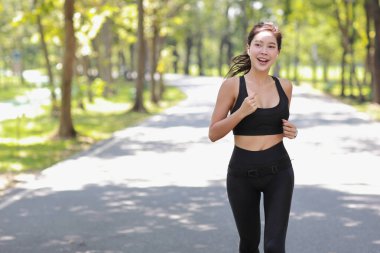 Güzel Asyalı fitness bayan sporcu. Sabah koşusu antrenmanı yapıyor. Açık havada. Parkta ağaçların altında koşan neşeli gülümseyen atlet kadın kameraya bakıyor ve müzik dinliyor.