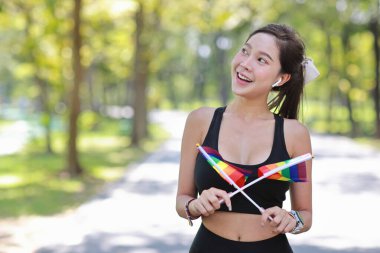 Genç neşeli Asyalı model transseksüel bileklikli LGBT gülümseyen ve kameraya bakarken gökkuşağı bayrağı sallıyor. Spor giyimli çekici kadın koşucunun fotoğrafı. gurur ve eşitlik kavramı.