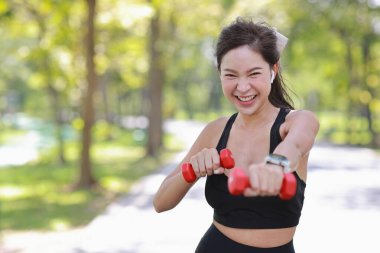 Güzel Asyalı fitness bayan sporcu sabah koşusundan sonra halter ağırlık kaldırıyor. Gülümseyen atlet kadın, parkta ağacın altında kameraya bakarken antrenman yapıyor.