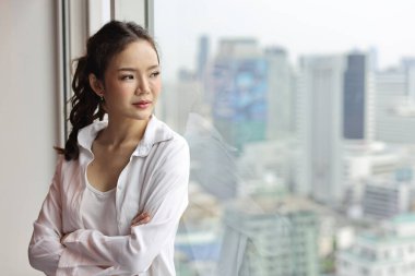 Kendine güvenen ve güzel Asyalı genç iş kadınının portresi mutlu gülümseyen yüzü ile modern bir otelde dikiliyor ve uzaklara bakıyor şehir arka planı bulanık. Başarı işi ve yaşam tarzı konsepti