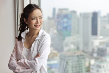 Kendine güvenen ve güzel Asyalı genç iş kadınının portresi mutlu gülümseyen yüzü ile modern bir otelde dikiliyor ve uzaklara bakıyor şehir arka planı bulanık. Başarı işi ve yaşam tarzı konsepti