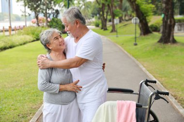 Asyalı son sınıf öğrencisi ya da bakıcı yaşlı bir adama parkta tekerlekli sandalyeyle yürümesine yardım ediyor. Yaşlı bir eş, evin dışında yaşlı bir kocaya iyi bakıyor ve destek oluyor..
