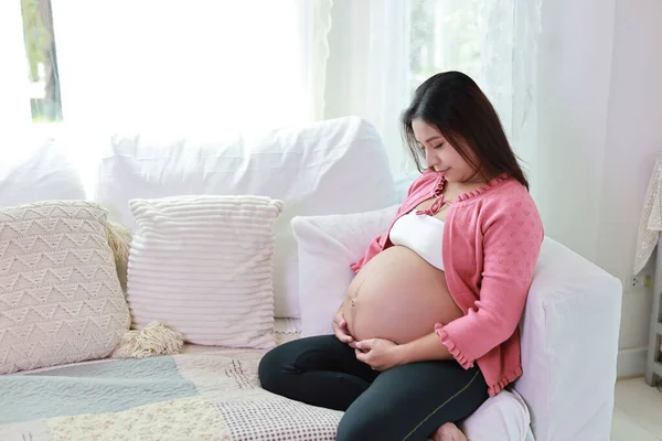 Happy smiling young asian pregnant woman sitting and resting on sofa in living room while touching and looking her belly. Expectant mother preparing and waiting for baby birth during pregnancy.