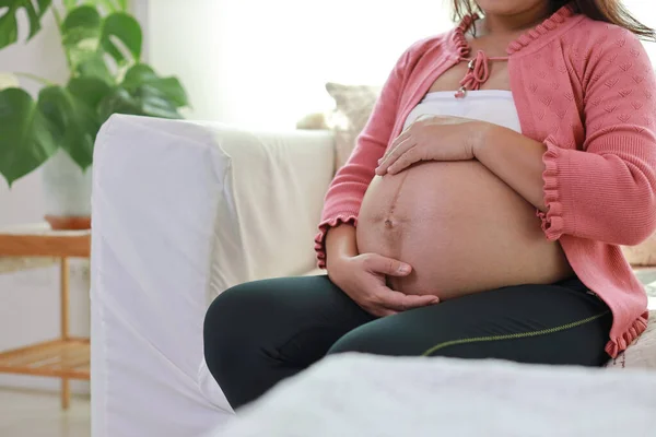 Happy young asian pregnant woman hands sitting and resting on sofa in living room while touching and looking her belly. Expectant mother preparing and waiting for baby birth during pregnancy.