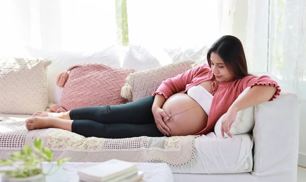 Happy smiling young asian pregnant woman resting and lying on sofa in living room while touching and looking at her belly. Expectant mother preparing and waiting for baby birth during pregnancy.