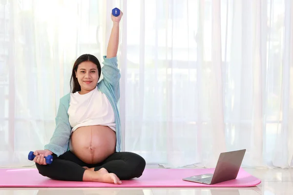 Happy young asian pregnant sit on pink mat lifting dumbbell and watch training video online from computer in living room at home. Expectant mother prepare for baby birth during pregnancy concept