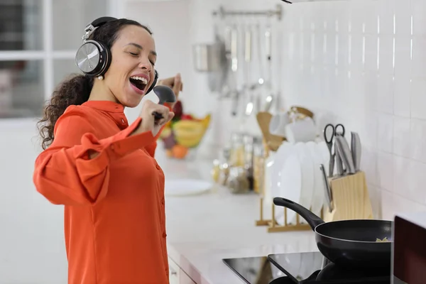 Happy latin woman cooking, smelling and tasting food while dancing in kitchen. Beautiful young female with headphone preparing delicious meal while listen music at home. Healthy and lifestyle concept