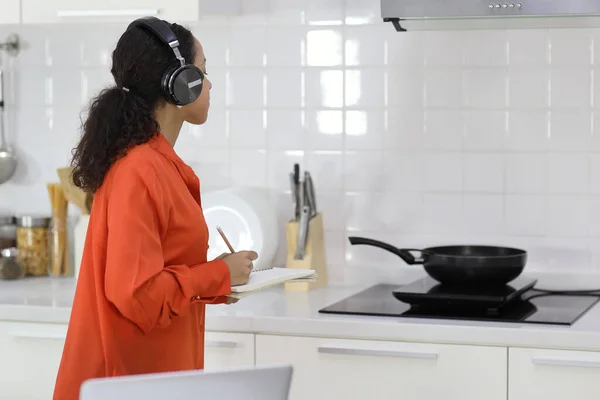 Rear view latin woman learn cooking food while check recipe from internet with computer in kitchen. Young female with headphone prepare delicious meal and listen music. Healthy and lifestyle concept
