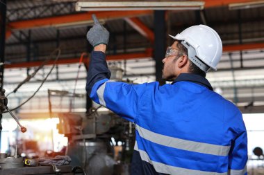 Arka görüş teknisyeni koruyucu giysi mühendisi ayakta ve telsiz kullanarak ağır sanayi imalat fabrikasında metal torna işletim makinesini kontrol ediyor veya onarıyor.