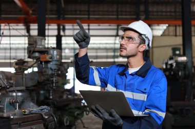 Mutlu teknisyen mühendis koruyucu kıyafetli adam bilgisayar kullanarak ağır sanayi imalat fabrikasında metal torna işi yapıyor.