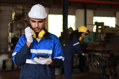 Koruyucu giysi standındaki teknisyen telsiz telsiz ve tablet kullanırken ağır sanayi imalat fabrikasındaki torna makinesini kontrol ya da bakım için çalıştırıyor.