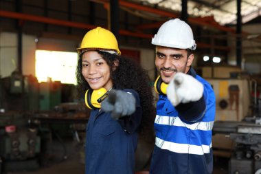 Koruyucu kıyafetli bir grup mühendis, ağır sanayi imalat fabrikasındaki metal torna makinesini kontrol ederken kameraya bakıyor ve işaret ediyor.