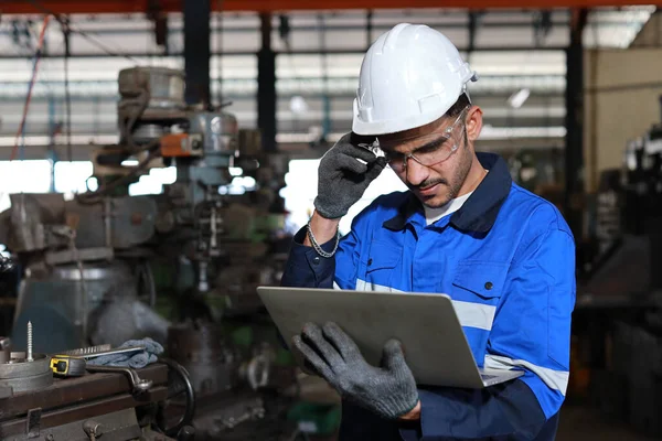 Teknisyen mühendis, ağır sanayi imalat fabrikasındaki metal torna işini kontrol ederken ayakta duran ve bilgisayar kullanan koruyucu giysiyi ciddi şekilde inceliyor.