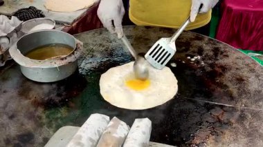 Bu resim, sıcak demir bir tawa üzerinde taze bir yumurtayı cızırdayan bir paratha üzerine kırarak lezzetli bir yumurta rulosu hazırlayan bir satıcıyı gösteriyor. Yakınlarda, güzelce paketlenmiş yumurta tepsileri, lezzet dolu, servis edilmeyi bekliyor..