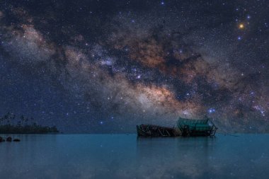 Ön planda yıkılmış bir balıkçı teknesi ve arka planda geceleri denizin üzerinde yükselen Samanyolu Galaksisi olan bir manzara fotoğrafı. Trat Eyaletinde çekilmiş, Koh Chang, Tayland.