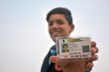 15-12-2021 Dewas, Madhya Pradesh, Hindistan. Koyu mavi okul üniforması giyen, gülümseyen yüzlü Hintli öğrenci elinde bulanık aadhar kartını gösteriyor. gökyüzü arkaplanı