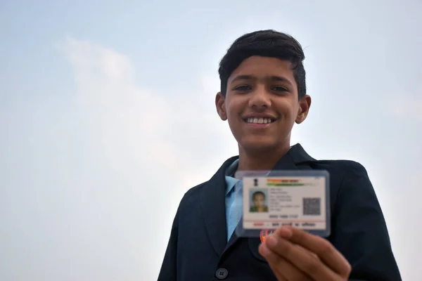 Indian school boy wearing dark blue school uniform, with smiling face shows her blurred aadhar card in her hand. sky background