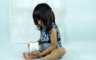 Beyaz arka planda oturan küçük bir çocuk ve elinde beyaz domuz kumbarası var. kaydetme kavramı.