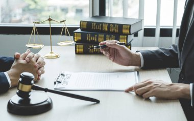 Avukatın eli kalem tutuyor ve ofisteki adama hukuki danışmanlık hizmeti veriyor. Adalet terazisi ve tokmak çekiciyle..