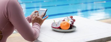 Meyve tepsisi, mayo giymiş Asyalı bir kadının eliyle otel havuzunun kenarında oturmuş online alışveriş için cep telefonunu kaydırıyor..