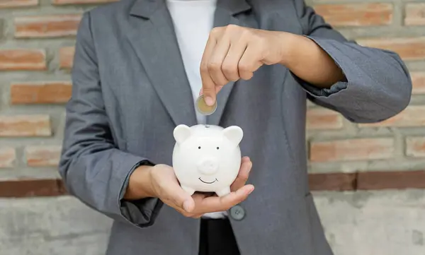 stock image Women hand in suit putting coins into white piggy bank, step up start-up business to success, Saving money for future plan and retirement fund concept.