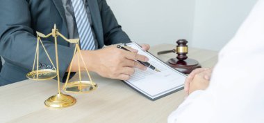 Avukatın eli kalem tutuyor ve ofiste hukuki danışmanlık hizmeti veriyor. Adalet terazisi ve tokmak çekiciyle..