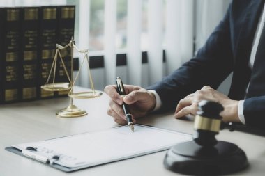Avukatın eli kalem tutuyor ve ofiste hukuki danışmanlık hizmeti veriyor. Adalet terazisi ve tokmak çekiciyle..
