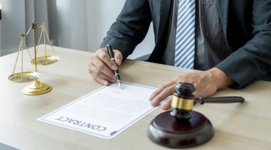 Avukatın eli kalem tutuyor ve ofiste hukuki danışmanlık hizmeti veriyor. Adalet terazisi ve tokmak çekiciyle..