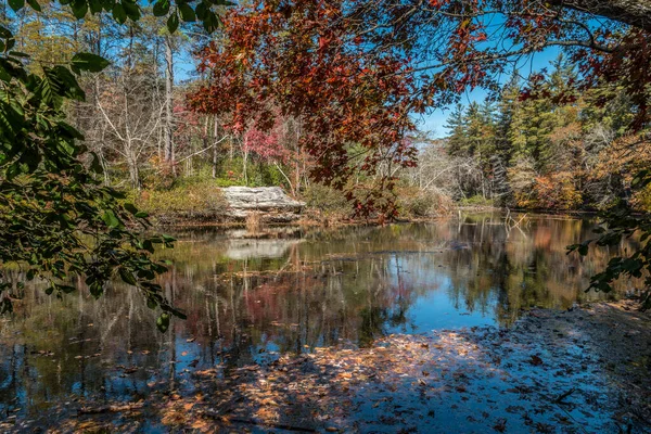 stock image Along the shoreline of the lake looking out through the trees at the colorful scene with mirrored reflection of the woodlands on the water and the falling leaves