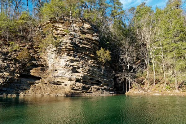 冬の晴れた日に川沿いの岩壁から小さな洞窟や木々が成長する崖に形成された岩の層 — ストック写真
