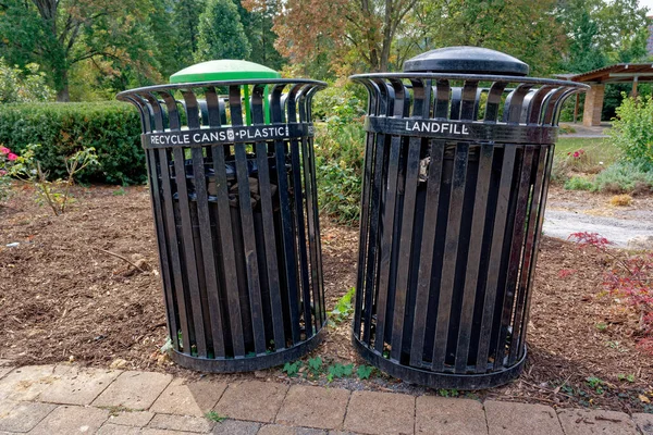 Two garbage cans along the walkway in the park one for cans and plastic bottles recyclables and the other is landfill garbage