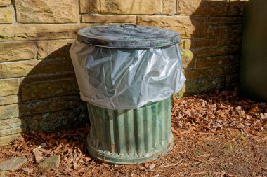 Soluk boyalı eski bir metal çöp tenekesi ve içinde beyaz bir plastik torba. Ayrıca güneşli bir günde açık hava binasına karşı bir kapak.