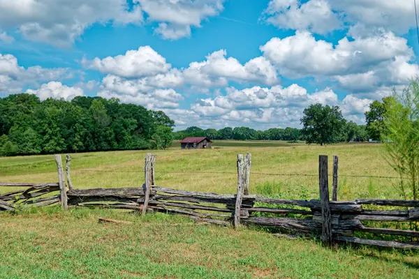 A hand built rustic wooden fence with barbwire around the farmland with an old barn in the background in the pasture on a sunny day with white fluffy clouds in late summertime