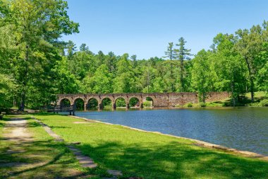 A landscape view of the walking path with stone benches and a fishing dock along the lake with the seven arch old stone bridge in the background on a bright sunny day in springtime clipart