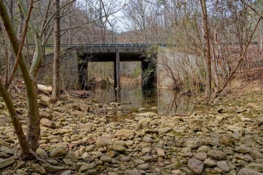 Korkuluğu olan büyük çimento bir viyadük yolu bir derenin üzerinden geçiyor. İlkbaharın başlarında ağaçlar ve çalılarla çevrili bir nehre taşan bir sürü kaya var.