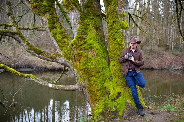 Young man stands leaning against a tree, wearing western wear and looking down at a camera in his hand; Bothell, Washington, United States of America