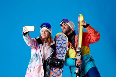 Kadın ve erkek kar kıyafetleriyle selfie çekiyorlar mavi arka planda kayak ve snowboardlarını gösteriyorlar..
