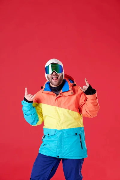 カメラを見ている間に彼の舌を突き出した雪の帽子 ジャケット ゴーグルの若い男の子と赤い背景に両手の指を上げて — ストック写真