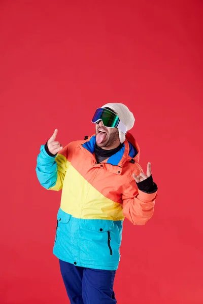 ジャケット 雪のゴーグルで彼の舌を突き出し 赤い背景に両手の指を上げて — ストック写真