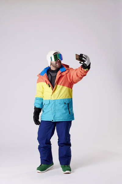 携帯電話で笑顔で白い背景で自撮りするスノーギアの青年 — ストック写真