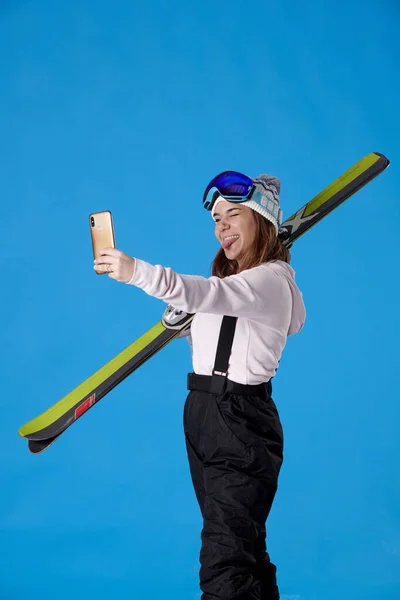 스키와 스노우 기어를 휴대폰을 여학생이 반짝이는 윙크를 파란색 배경에 내밀고 — 스톡 사진
