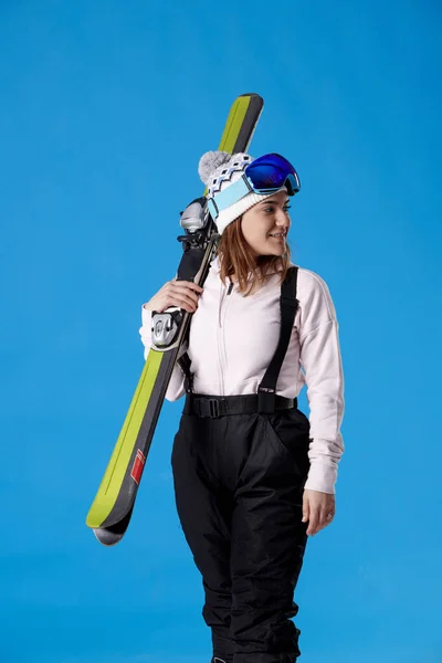 雪のギアを身に着けている笑顔の女性は 側面を見て 青い背景にスキーのペアを保持 — ストック写真