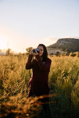 Genç bir yetişkin, antika kamerasıyla çayırda yürüyor, günbatımının keyfini çıkarıyor ve fotoğraf çekiyor..