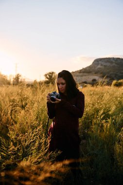 Genç bir yetişkin, antika kamerasıyla çayırda yürüyor, günbatımının keyfini çıkarıyor ve fotoğraf çekiyor..