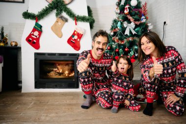 Bir aile, baba, anne ve kız, aynı Noel pijamalarını giyiyorlar, şöminenin önünde aile fotoğrafları çekiyorlar ve bir Noel ağacı. Sahne, tatil sezonunun neşesini, sevgisini ve birlikteliğini temsil ediyor.