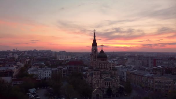 夕方の紫とオレンジの夕日の景色の雲と聖降誕大聖堂から後方に飛ぶ 空中ビューウクライナのダウンタウンでハリコフ市正統派教会 — ストック動画