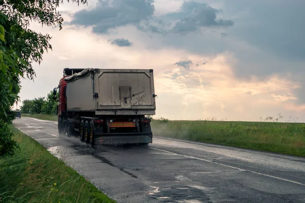 stock image Heavy truck vehicle driving wet rural asphalt road in rainy weather with epic clouds and green landscape. Rear view of the car with water mist on wheels