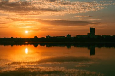 Gün doğumu güneşi gölün üzerinde parlıyor. Bulutlar sakin ayna su yüzeyine yansıyor. Şehir parkında, çok katlı binalarla dolu renkli bir sabah.