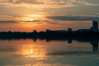 Bulutlu gökyüzünde, göl suyuna yansıyan güneşli bir gün doğumu. Nehir kenarında huzurlu bir sabah